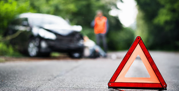 Personne avec un gilet rouge à côté d'une voiture accidentée avec un triangle de signalisation sur la chaussée