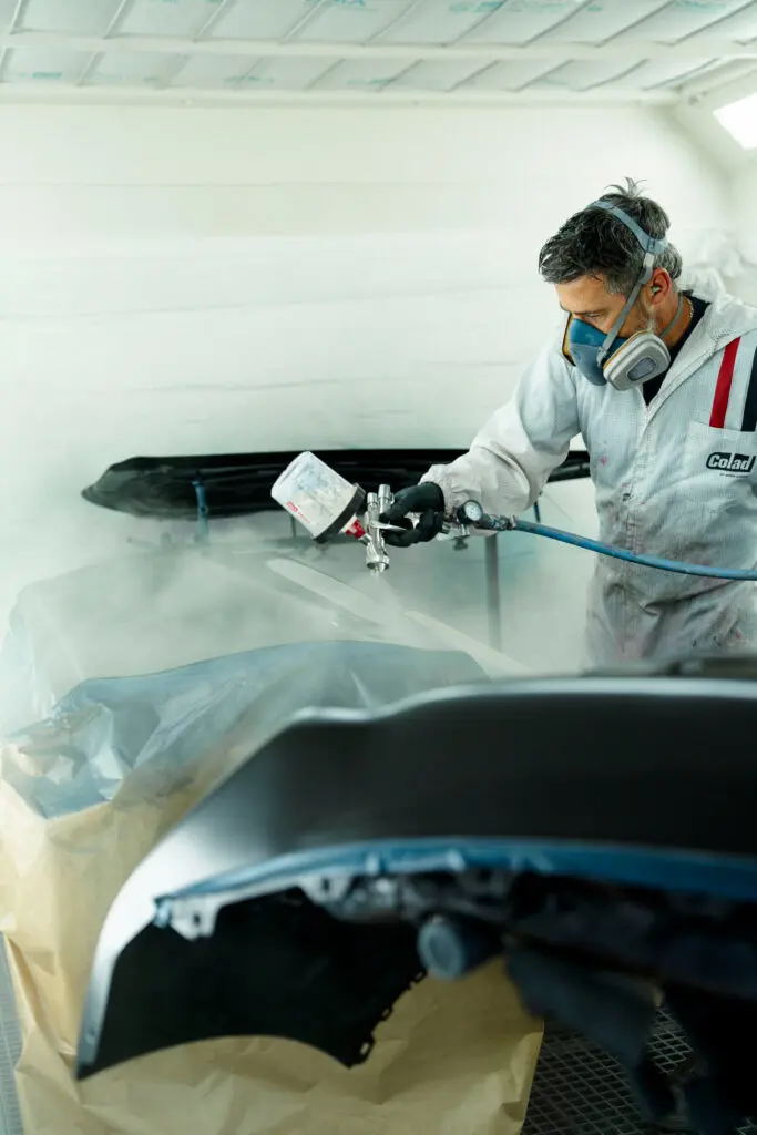 Homme en combinaison de peinture portant également un masque de peinture entrain de peindre un élément de carrosserie dans une cabine de peinture