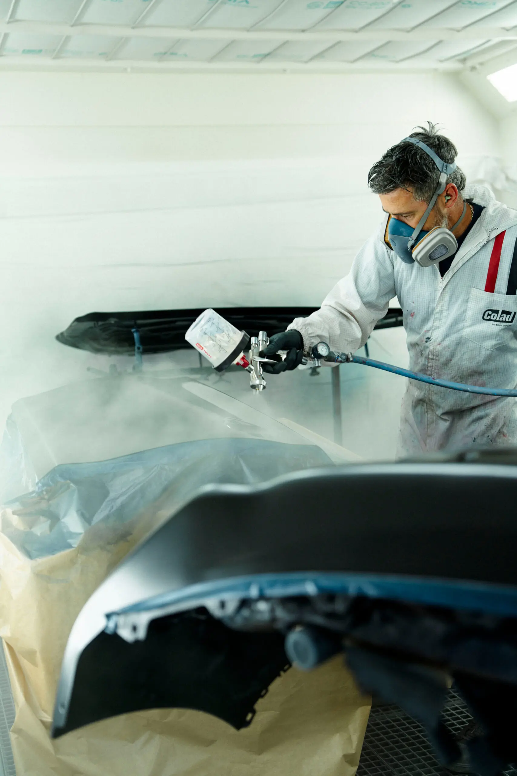 Homme en combinaison de peinture portant également un masque de peinture entrain de peindre un élément de carrosserie dans une cabine de peinture