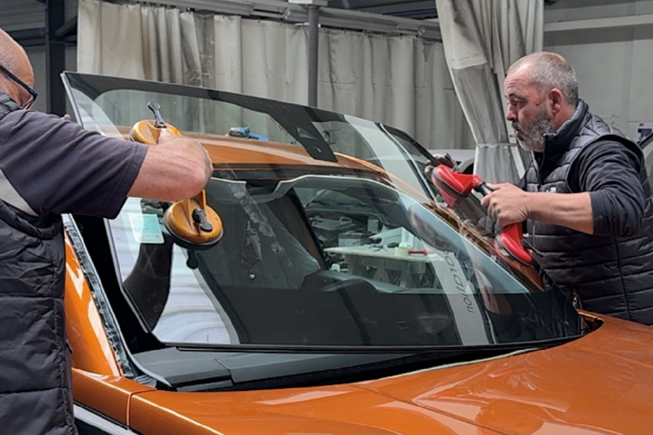 Photo de deux carrossiers en train de déposer un pare-brise sur une voiture de couleur orange.